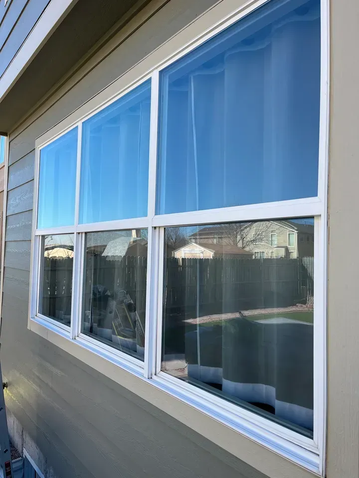 Person cleaning a house window with a squeegee, standing beside a beige exterior wall at sunset.