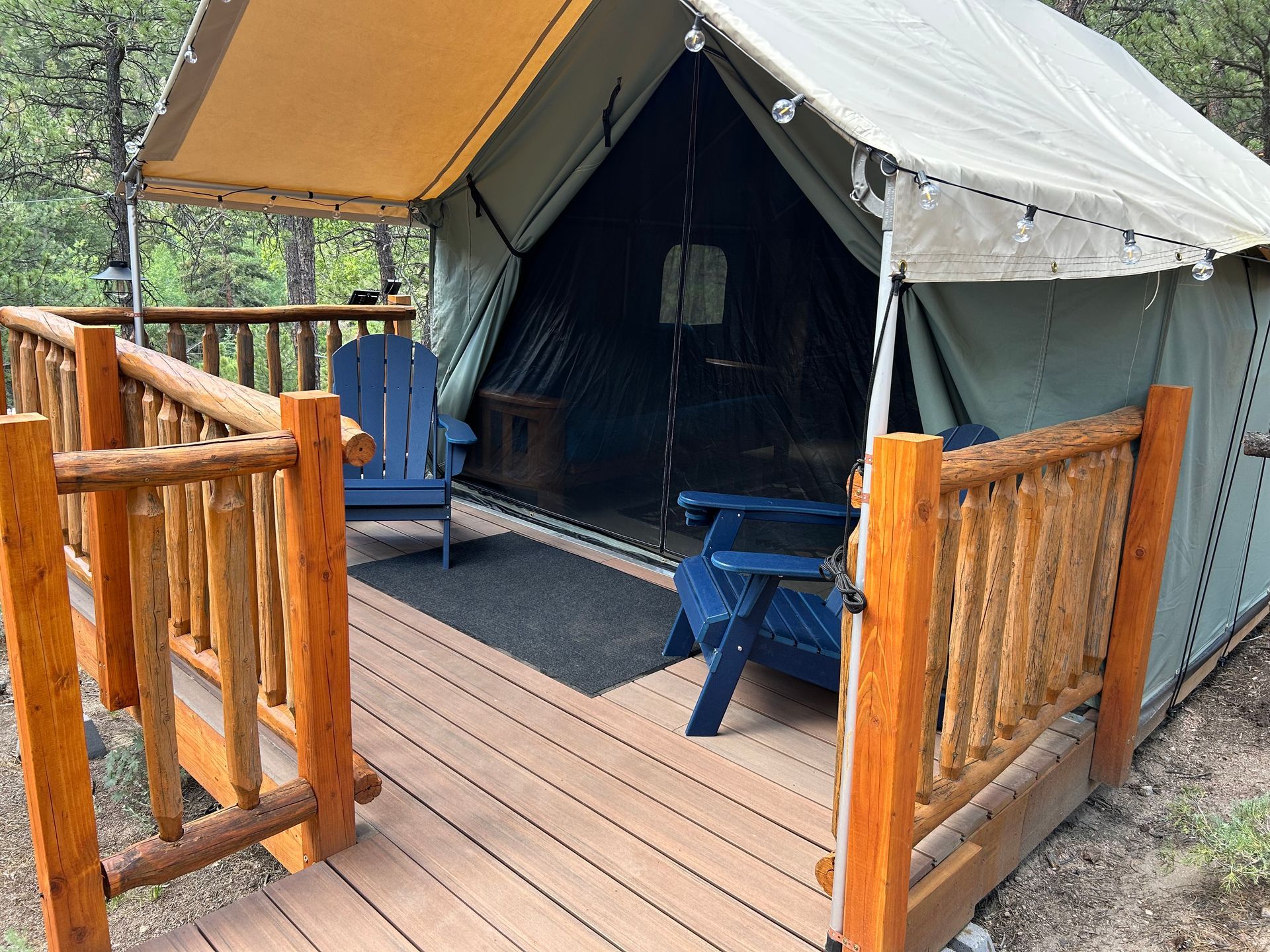 A glamping tent with a wooden deck and railing, two blue chairs, and a canopy.
