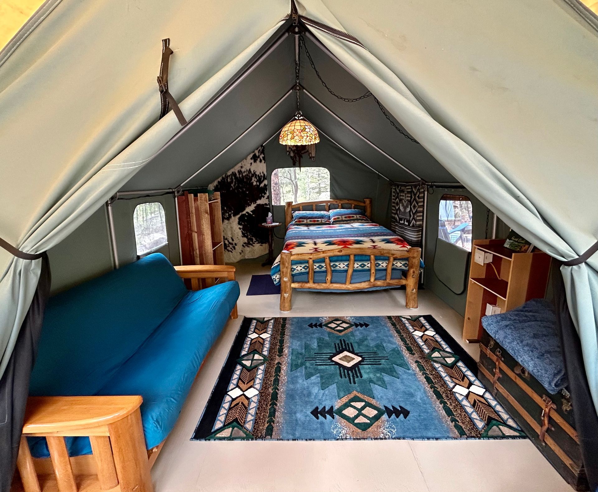 Interior of a glamping tent with bed, sofa, rug, wooden furniture, and decorative lighting.