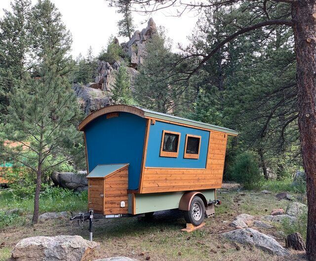 Tiny wooden cabin on wheels in a wooded area, with blue and brown exterior.