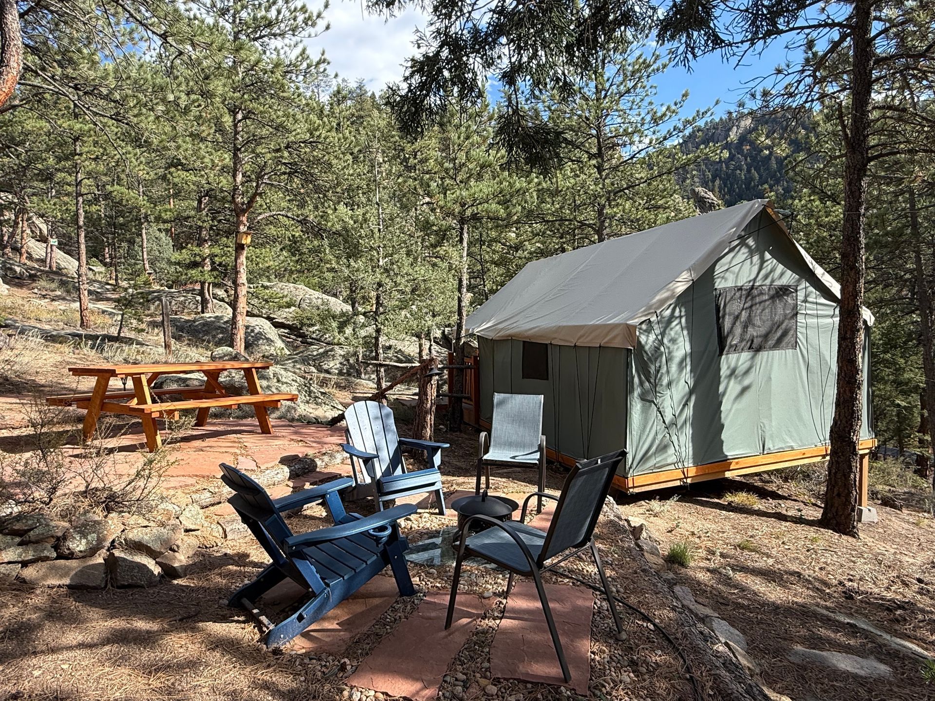 Camping glamping site with a tent, seating, and picnic table in a wooded area.