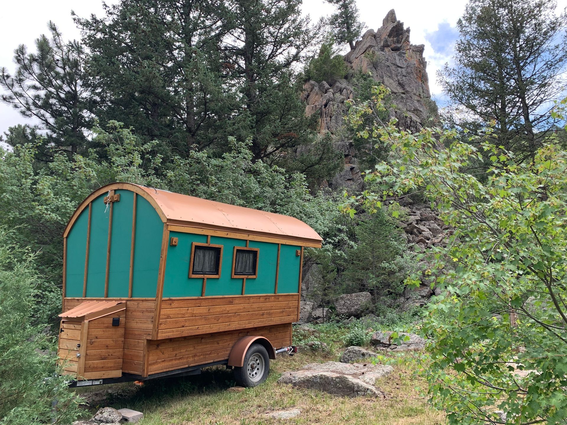 Wooden travel trailer with teal roof and wooden siding, parked near a rocky hillside and trees.