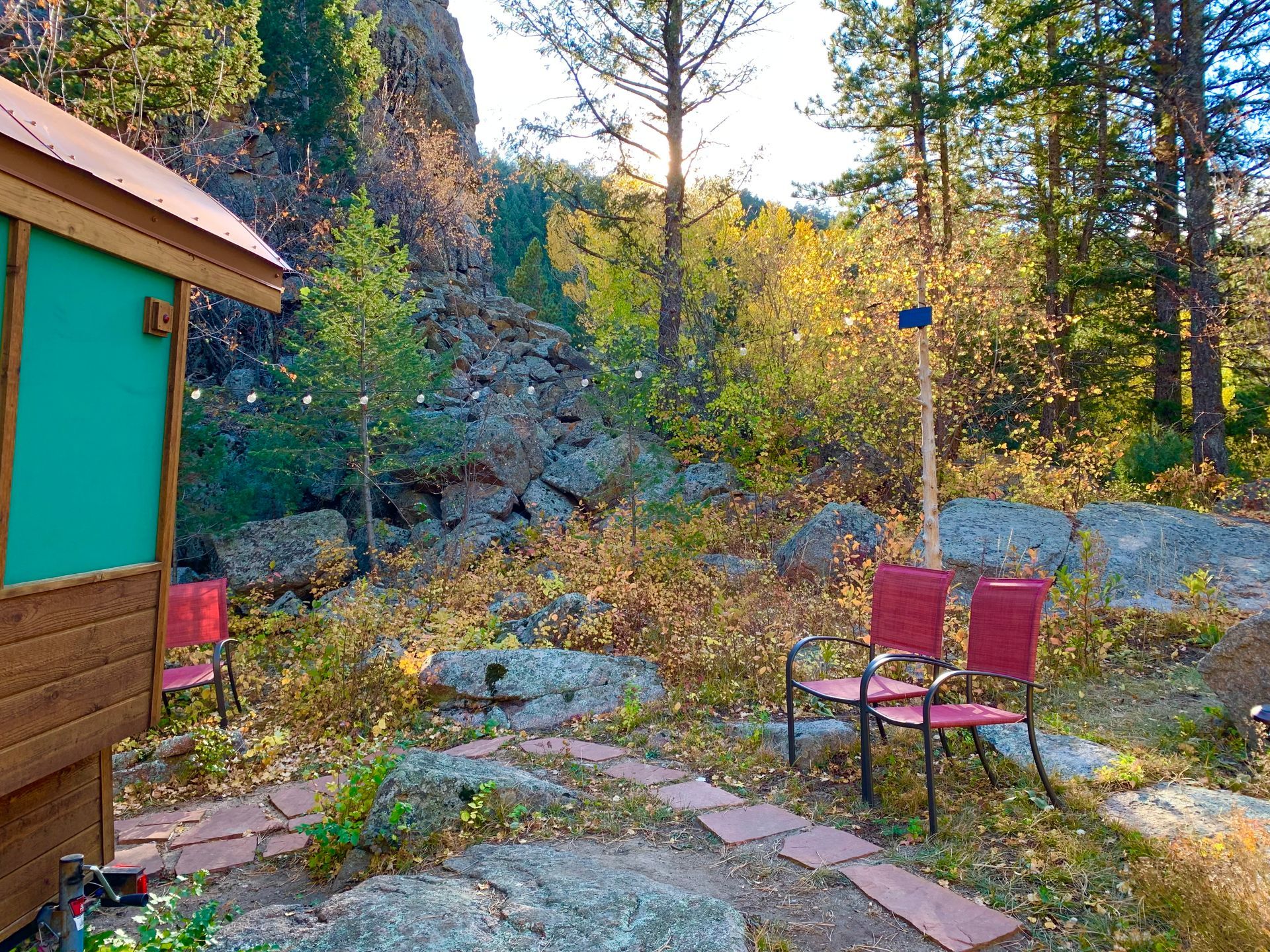 Red chairs on a stone path beside a cabin with natural landscape, trees, and rocks in sunlight.