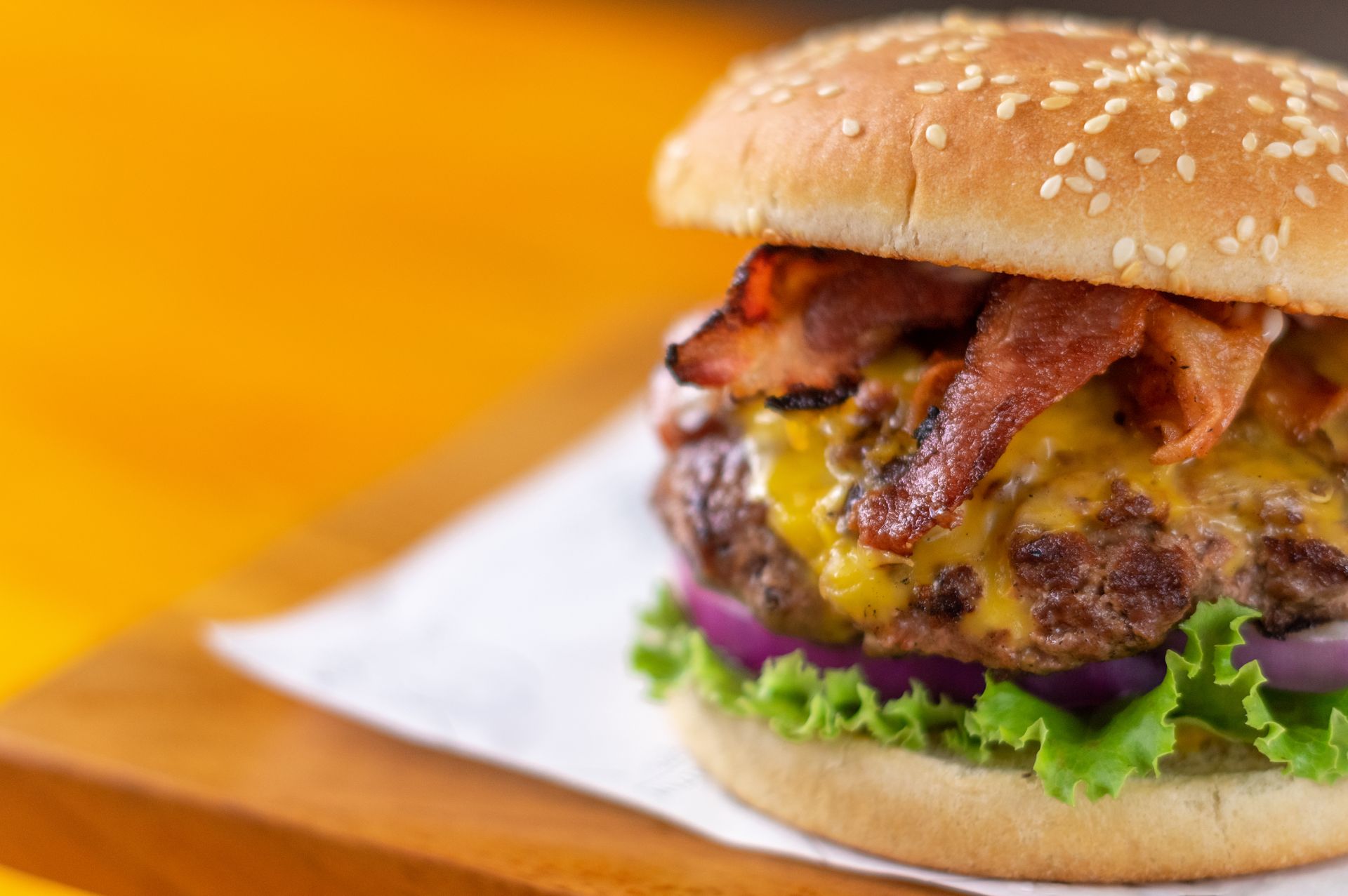Burger with bacon, cheese, lettuce, and red onion on a sesame seed bun, on a wooden board.
