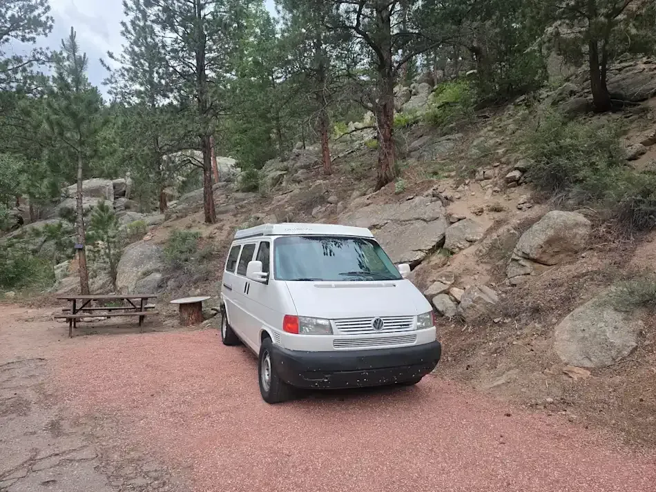 White camper van parked in a rocky campground, trees in the background, cloudy sky.