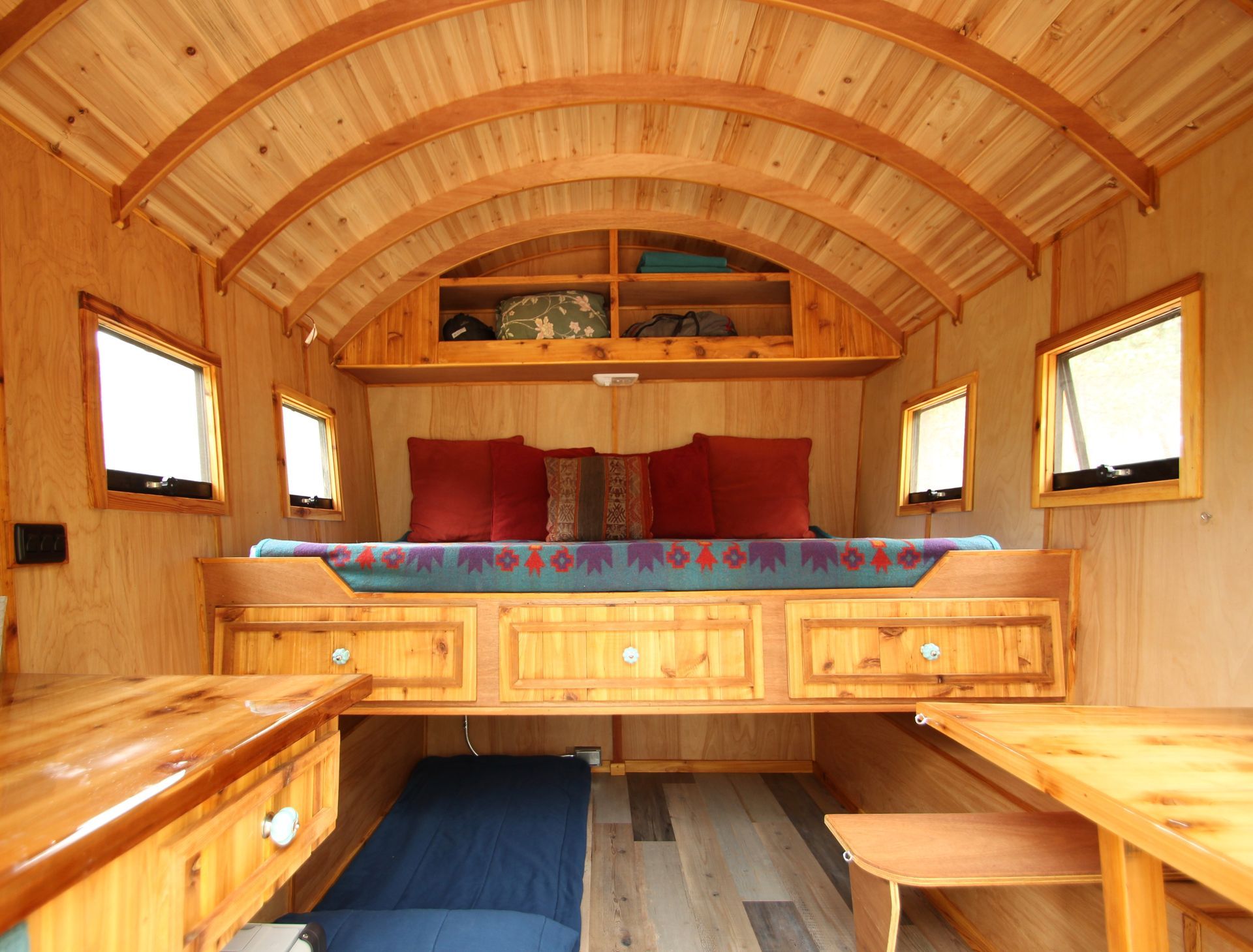 Wooden interior of a small home with bed, windows, and storage.  Natural wood tones.