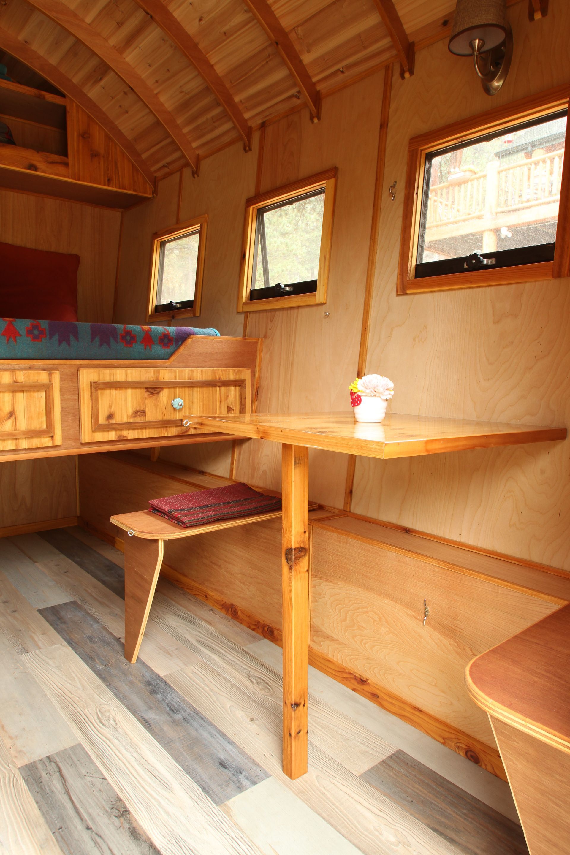 Interior view of a small wooden cabin with a table, bench, windows, and bed.