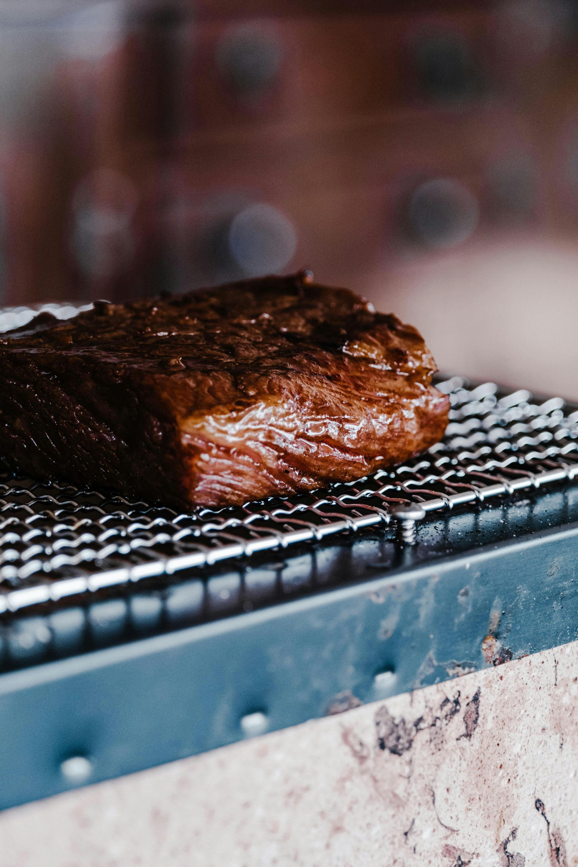 Large, seared piece of meat on a metal grill. Exterior is dark brown, interior is red, with a blurry background.