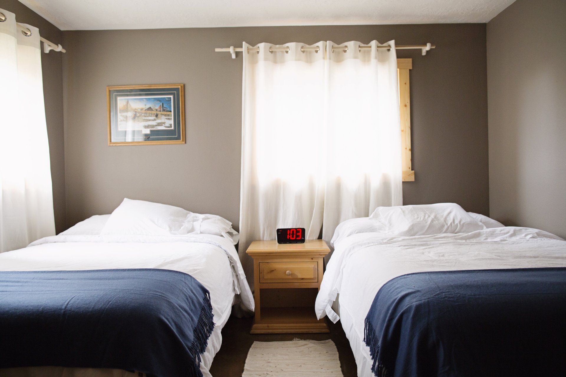 Twin beds in a small bedroom with dark blue blankets, a window with white curtains, and a nightstand between them