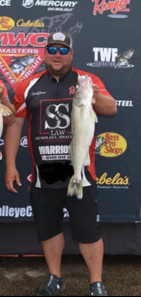 Man holding a large fish on a tournament stage, wearing sponsor gear and sunglasses