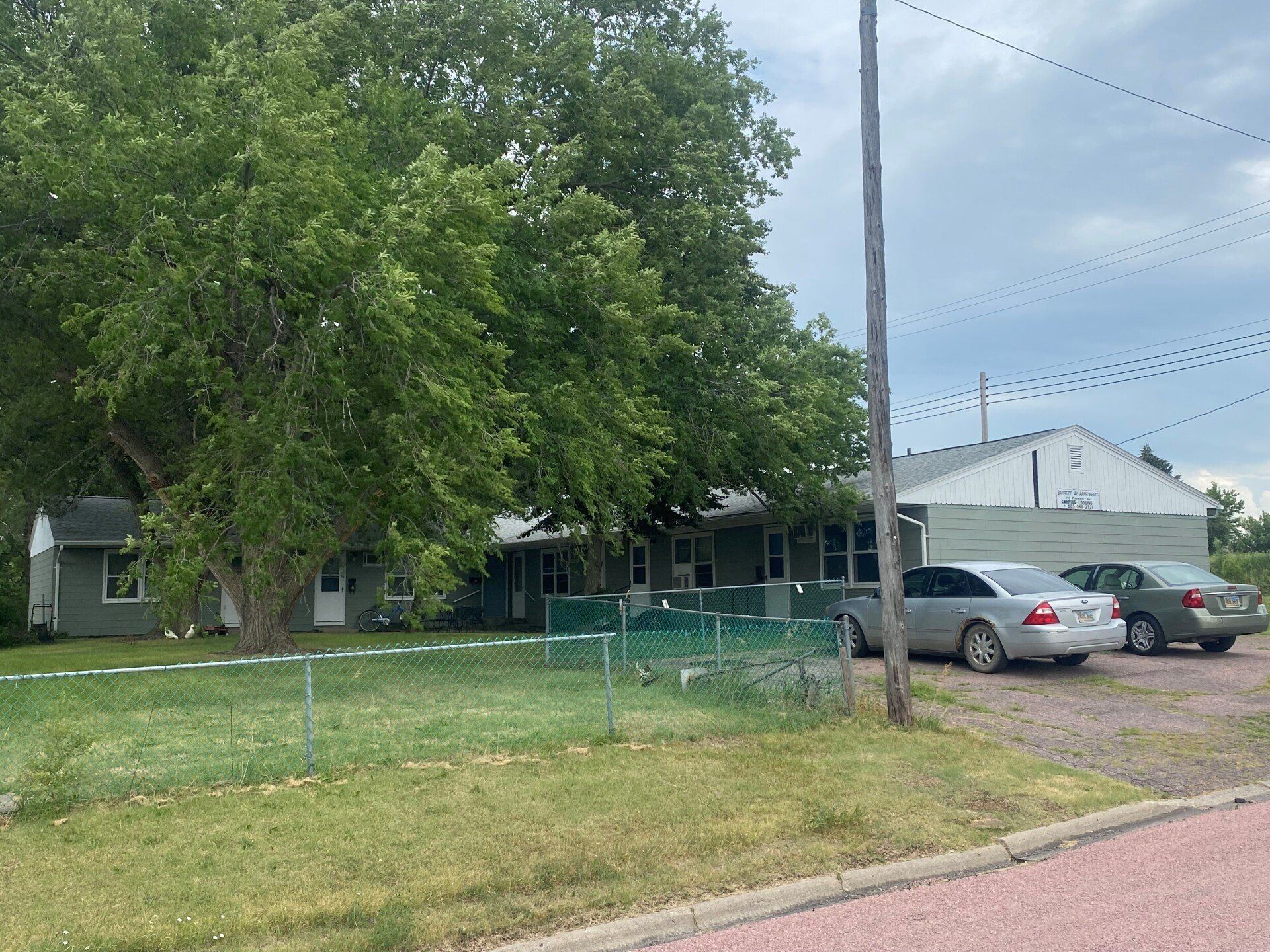 Suburban house with large tree, green lawn, chain-link fence, and two parked cars by the driveway