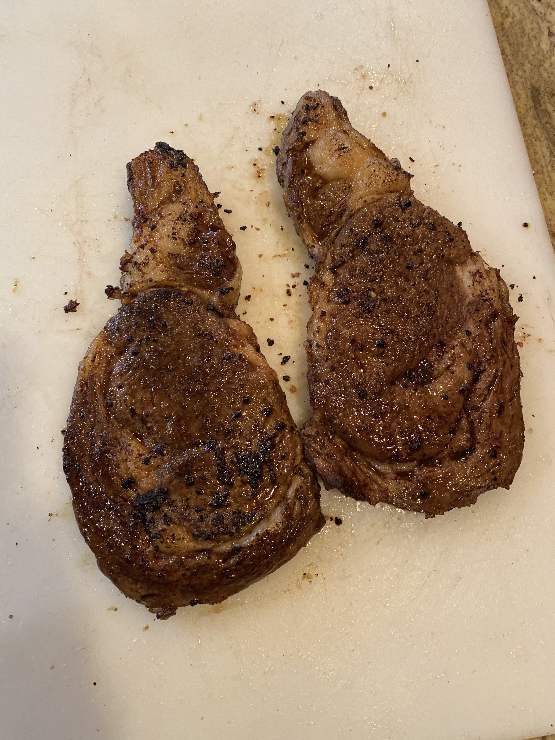 Two browned meat pieces on a white cutting board, with seasoning scattered around them