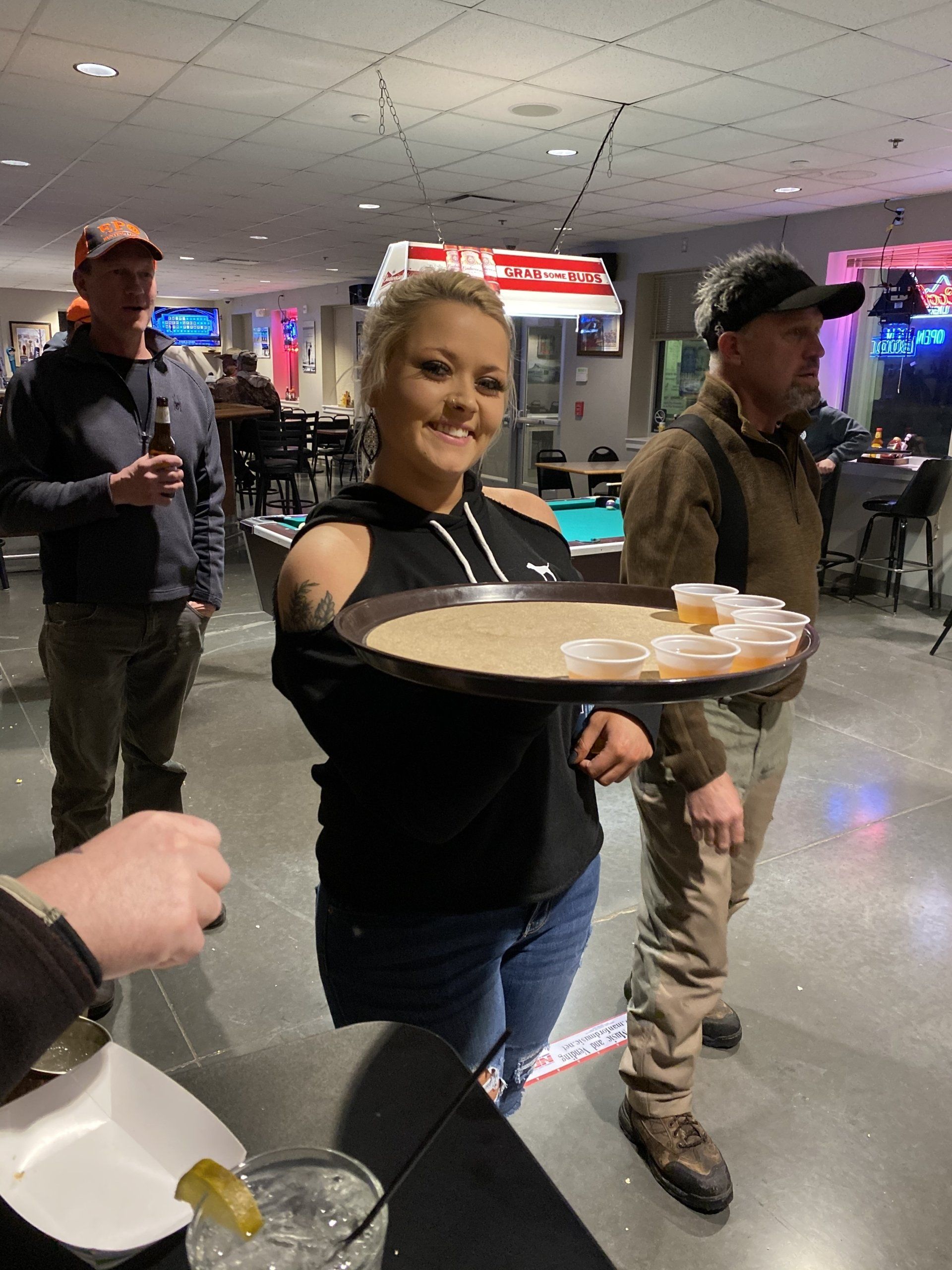 Woman in a black top carrying a tray of beer samples in a hall while people stand nearby