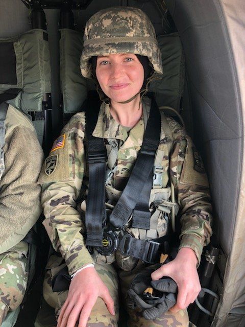 Soldier in camouflage uniform and helmet seated inside a military aircraft, wearing a harness and smiling.