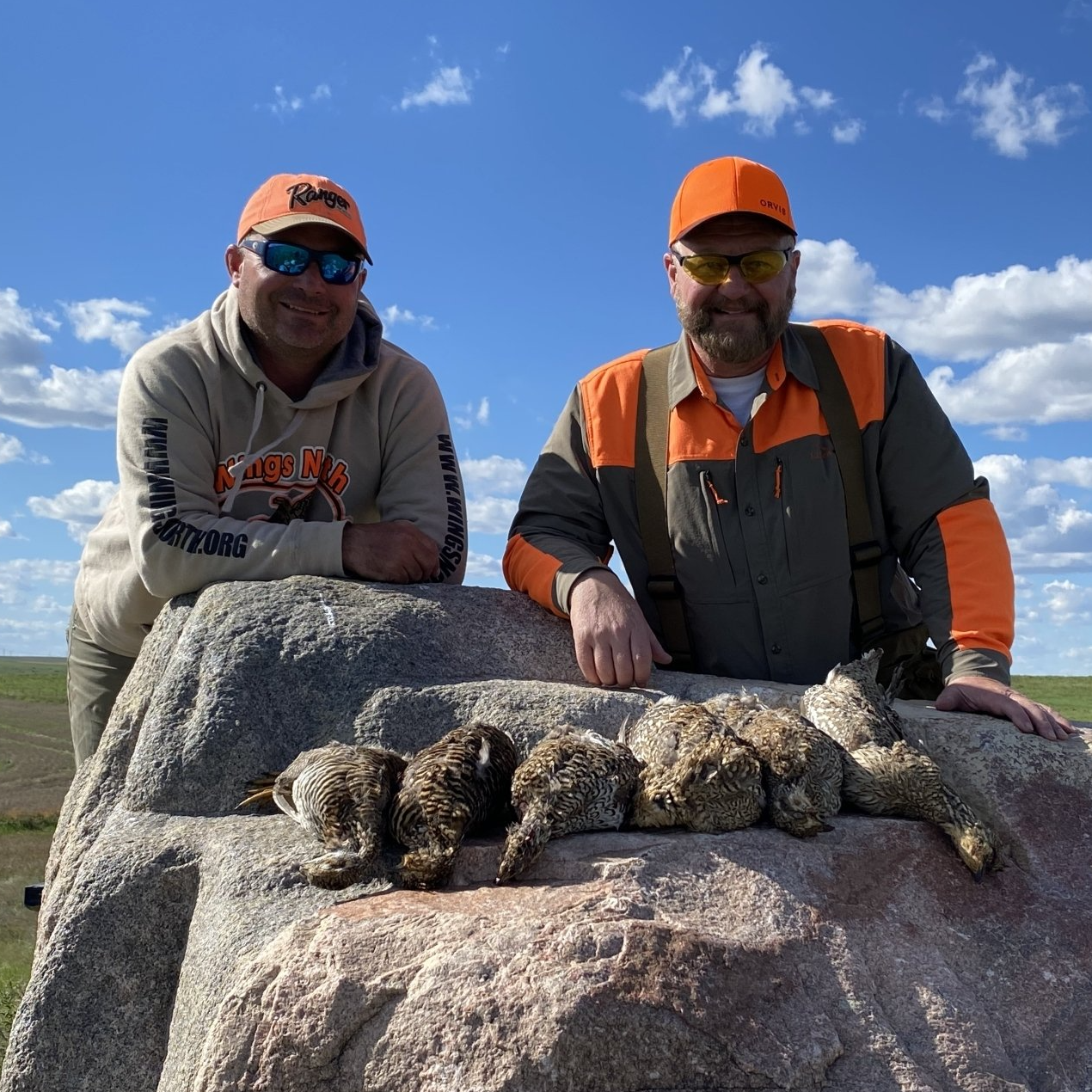Two hunters in orange caps pose behind a big game animal on a rock under a blue sky.