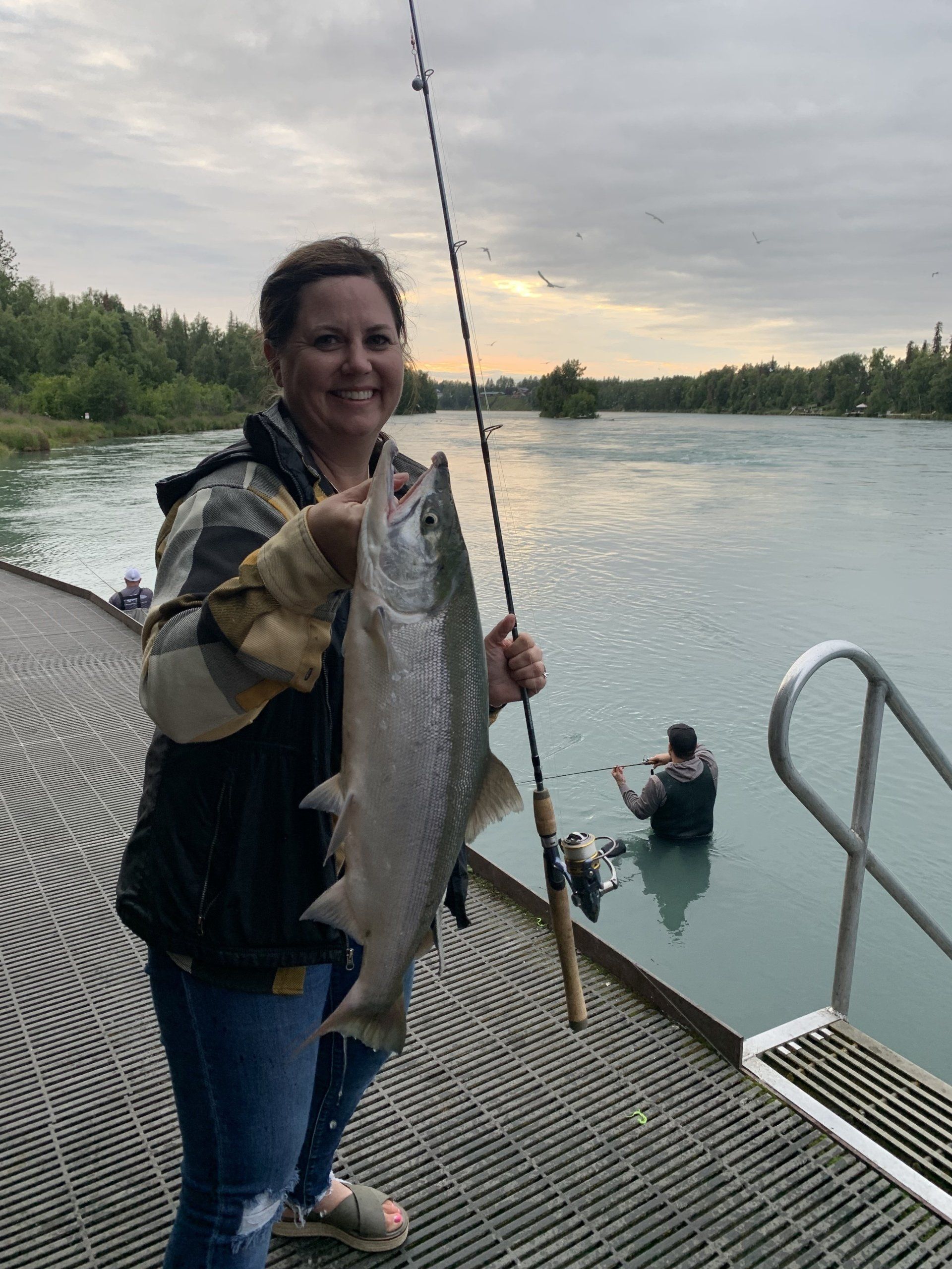 Person on a riverside dock holds a large fish, with another person in the water nearby at sunset