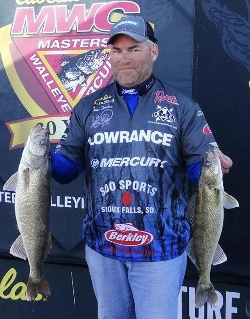 Angler in blue sponsor jersey holding a large fish at a tournament podium with MWC backdrop