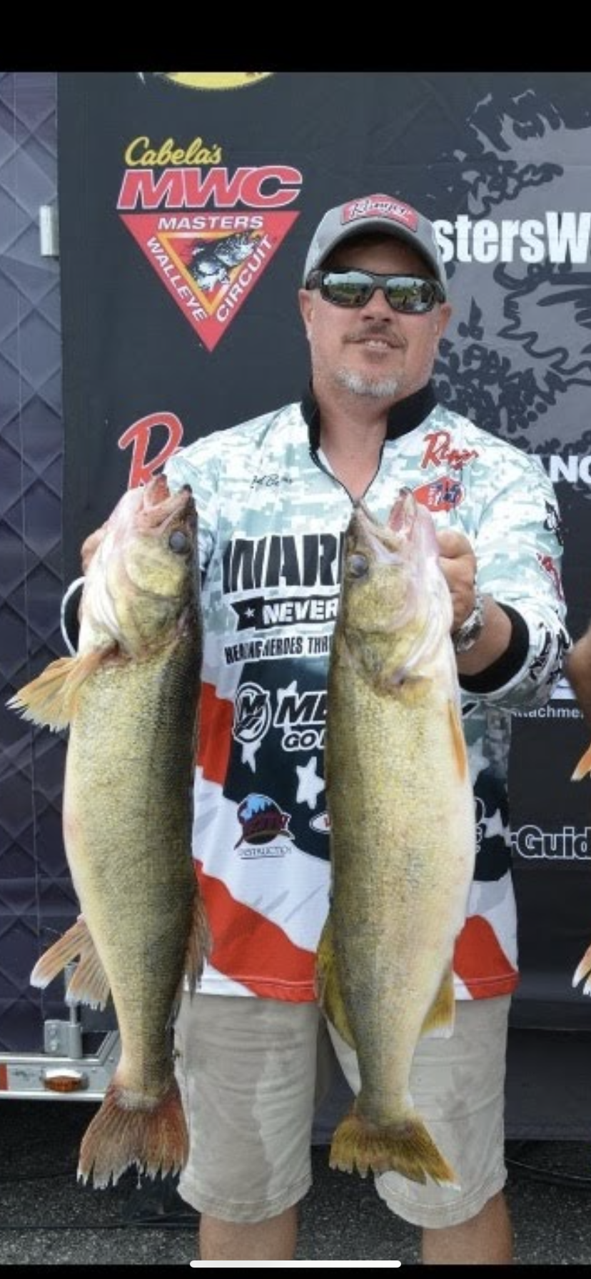Man holding two large fish at an MWC fishing event, wearing sunglasses and a white sponsor jersey.