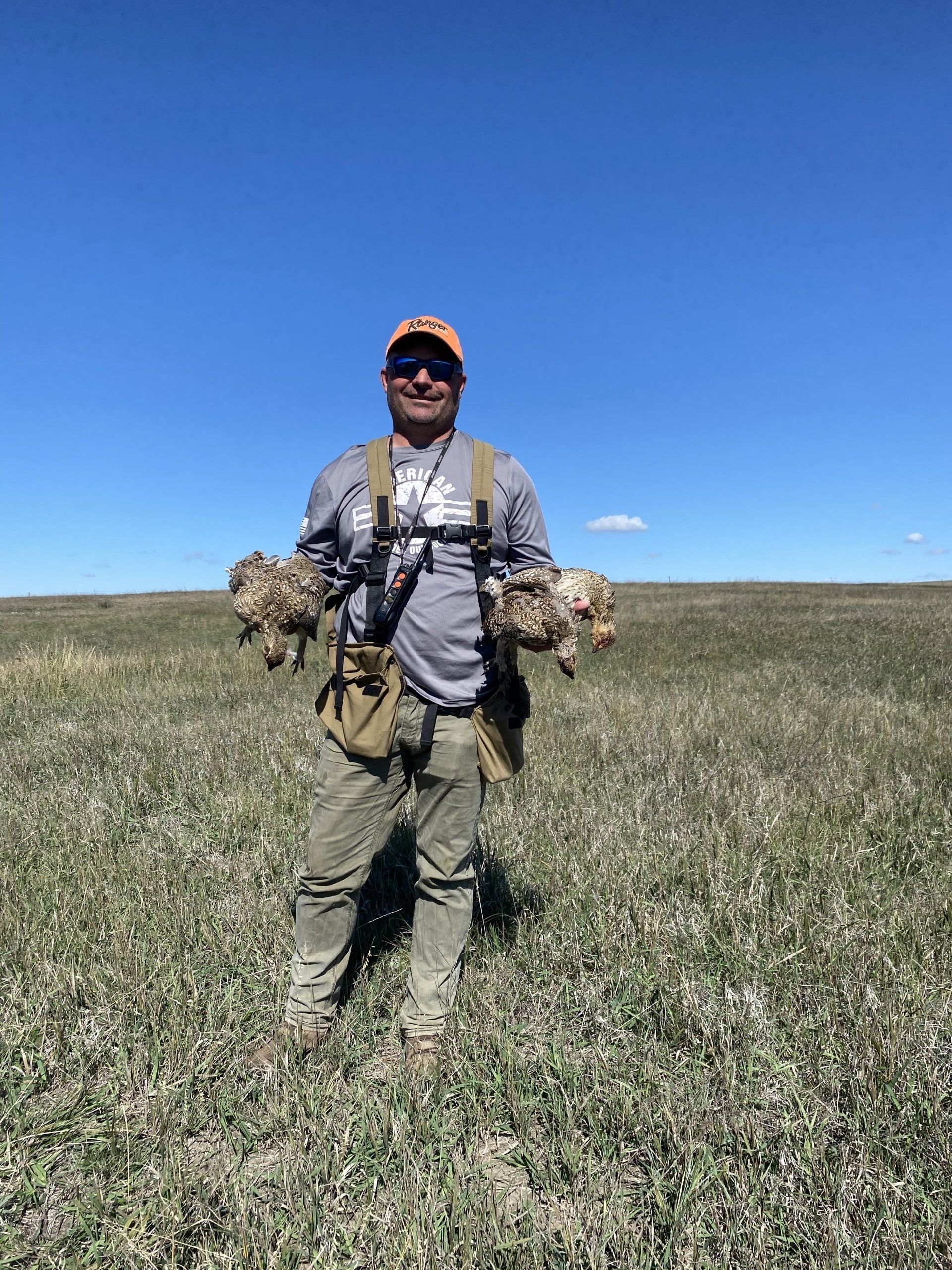 Person standing in a grassy field holding game birds, under a clear blue sky.