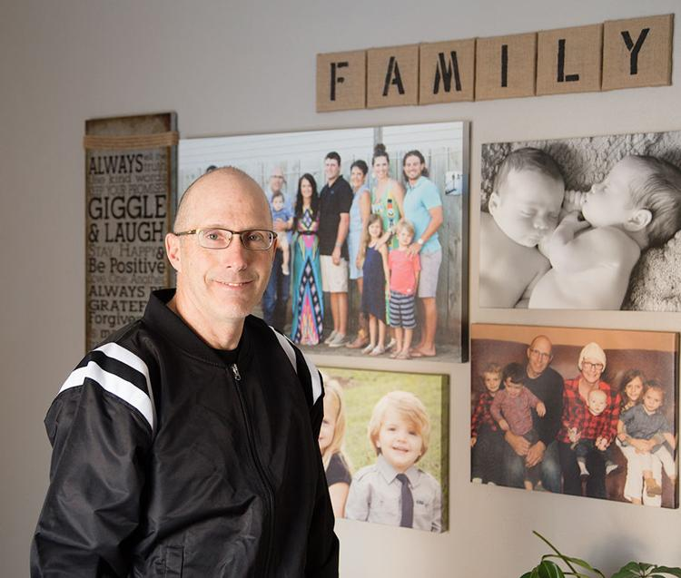 Man standing beside a wall of family photos and the word “FAMILY” displayed above them.