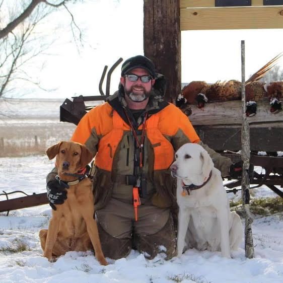 Man in orange jacket with two dogs in the snow beside a wooden shelter