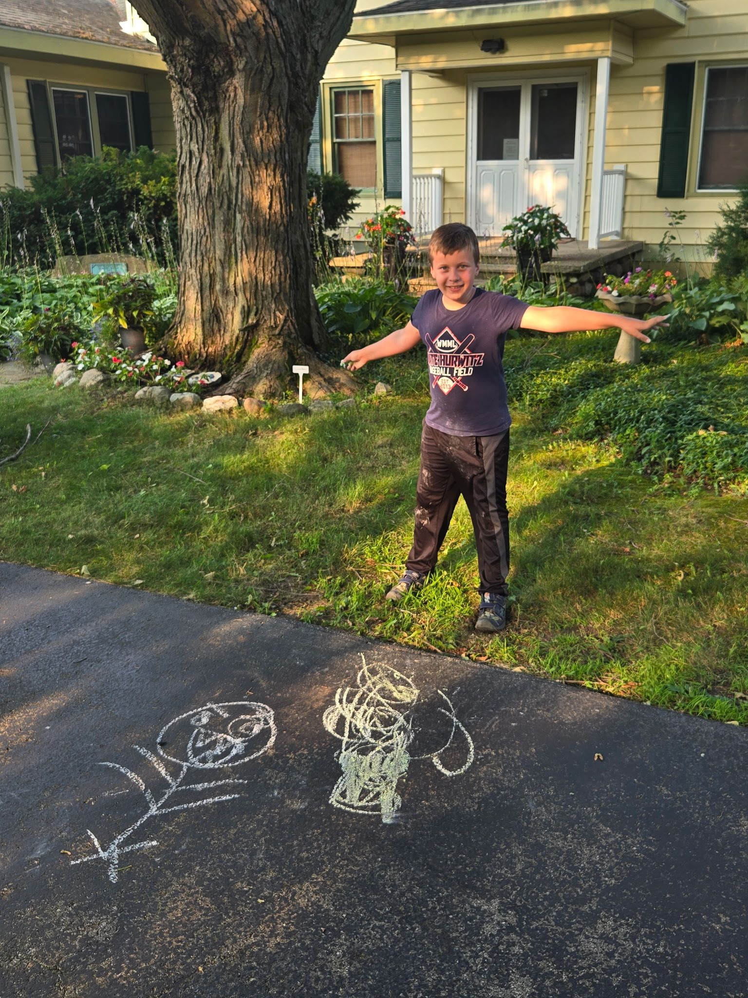 Boy standing in front of a house, arms outstretched, near chalk drawings on asphalt.