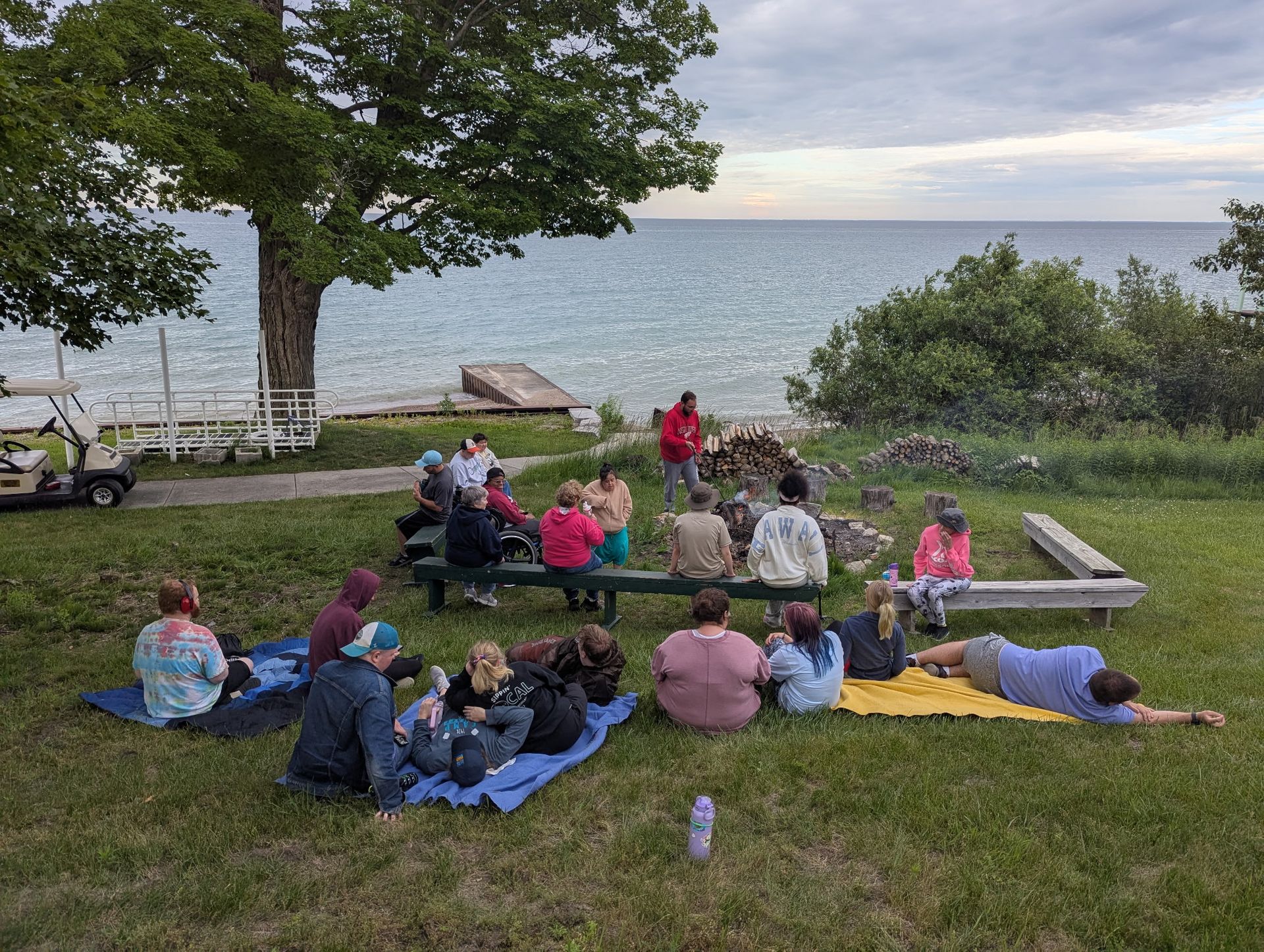 People sitting around a campfire on a grassy area near a lake. Dusk sky, trees and a golf cart are in view.