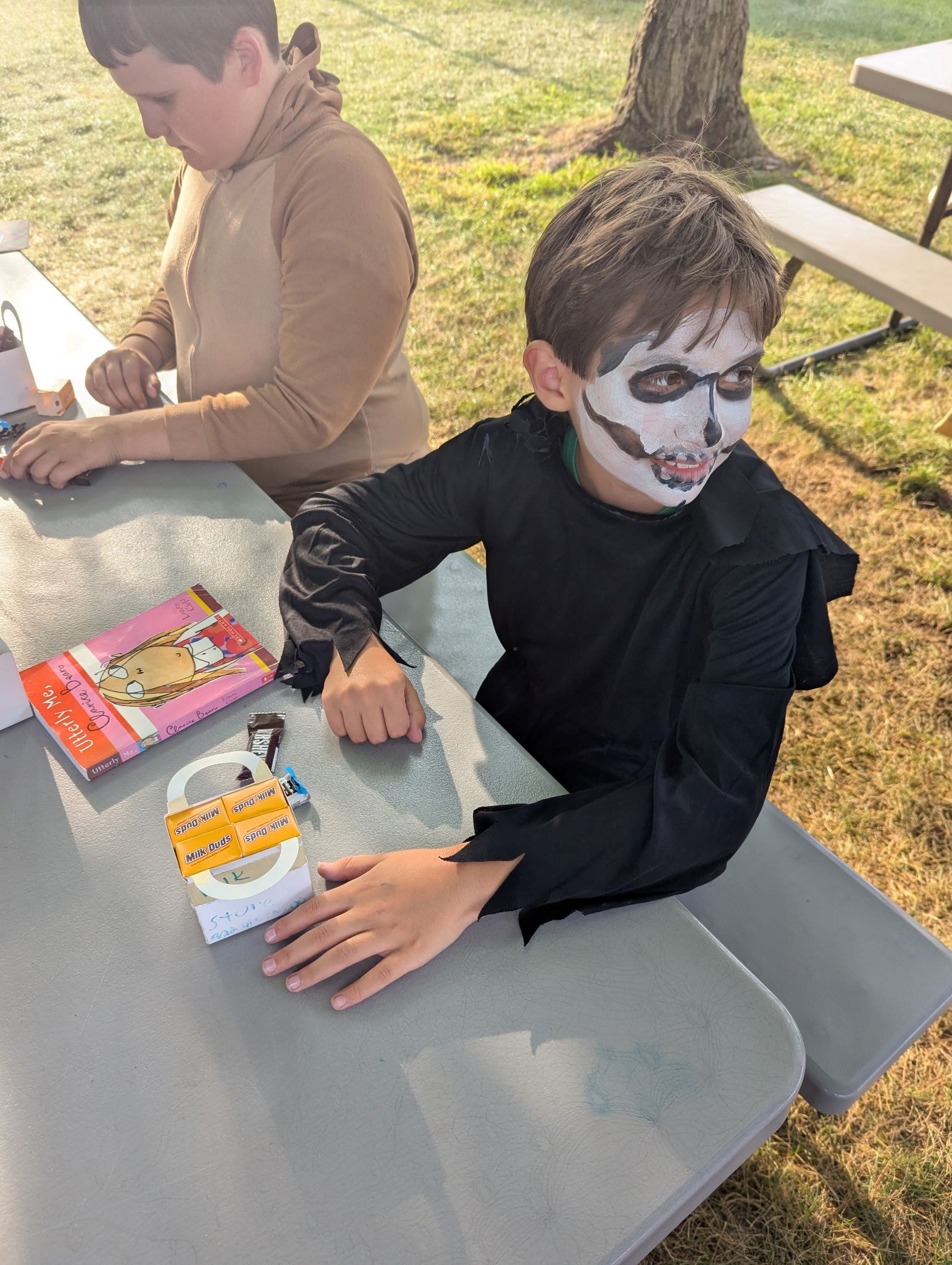 Boy in skull face paint and black costume at a picnic table, crafting. Another person in background.