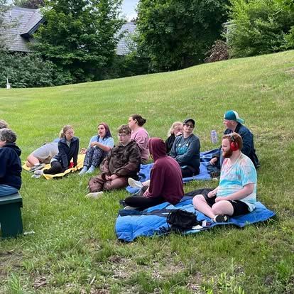 People sitting on a grassy hill, listening. Some have blankets, wearing casual clothing.