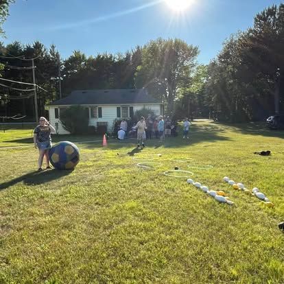 People playing games in a sunny yard. House in the background. Large ball and bowling pins on grass.