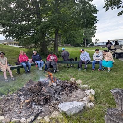 People gathered around a campfire on grass, sitting on a bench. Cloudy day, with golf course in background.
