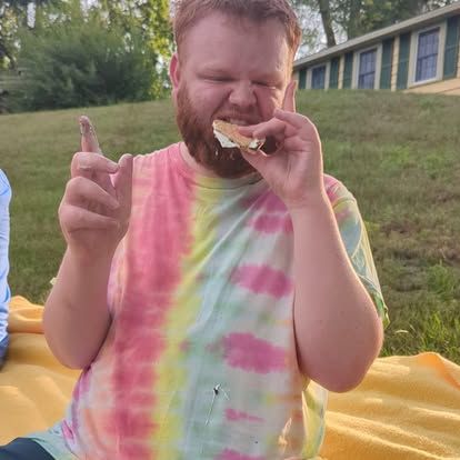 Man in tie-dye shirt eats s'more outdoors, pointing finger. Green grass, yellow blanket, house in background.