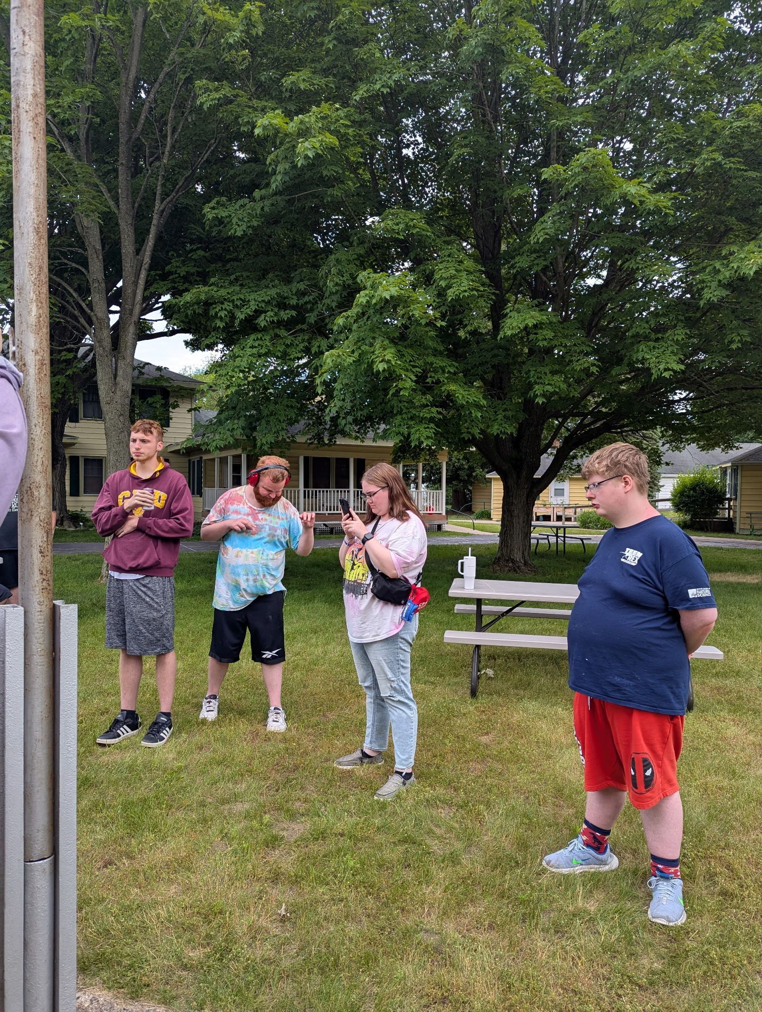Four people stand outside on grass near a tree. One in red shorts and a blue shirt looks on.