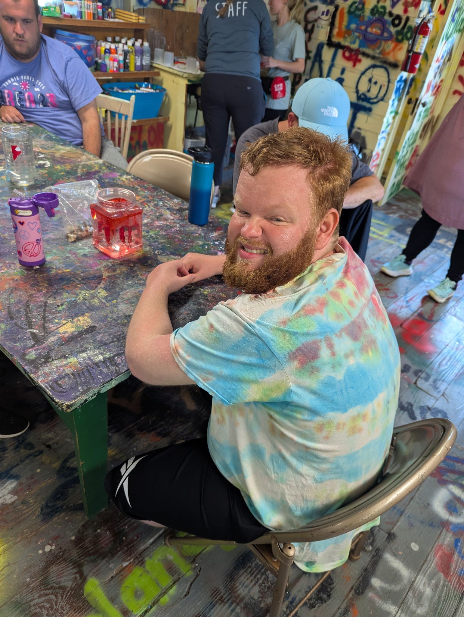 Man with a red beard smiles in a tie-dye shirt at a craft table in a colorful room.