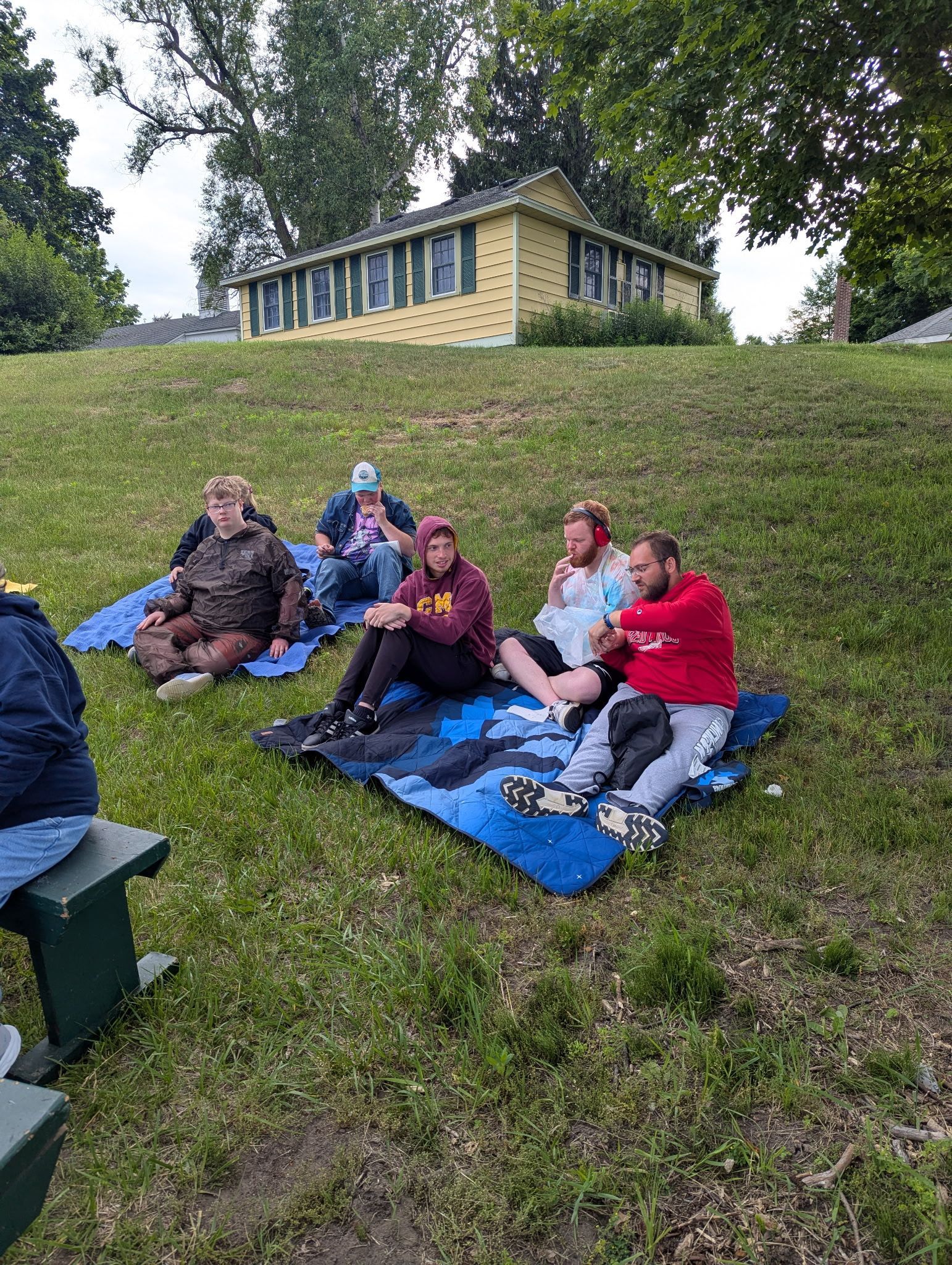 People sitting on blankets on a grassy hill, watching something. Yellow house in the background.