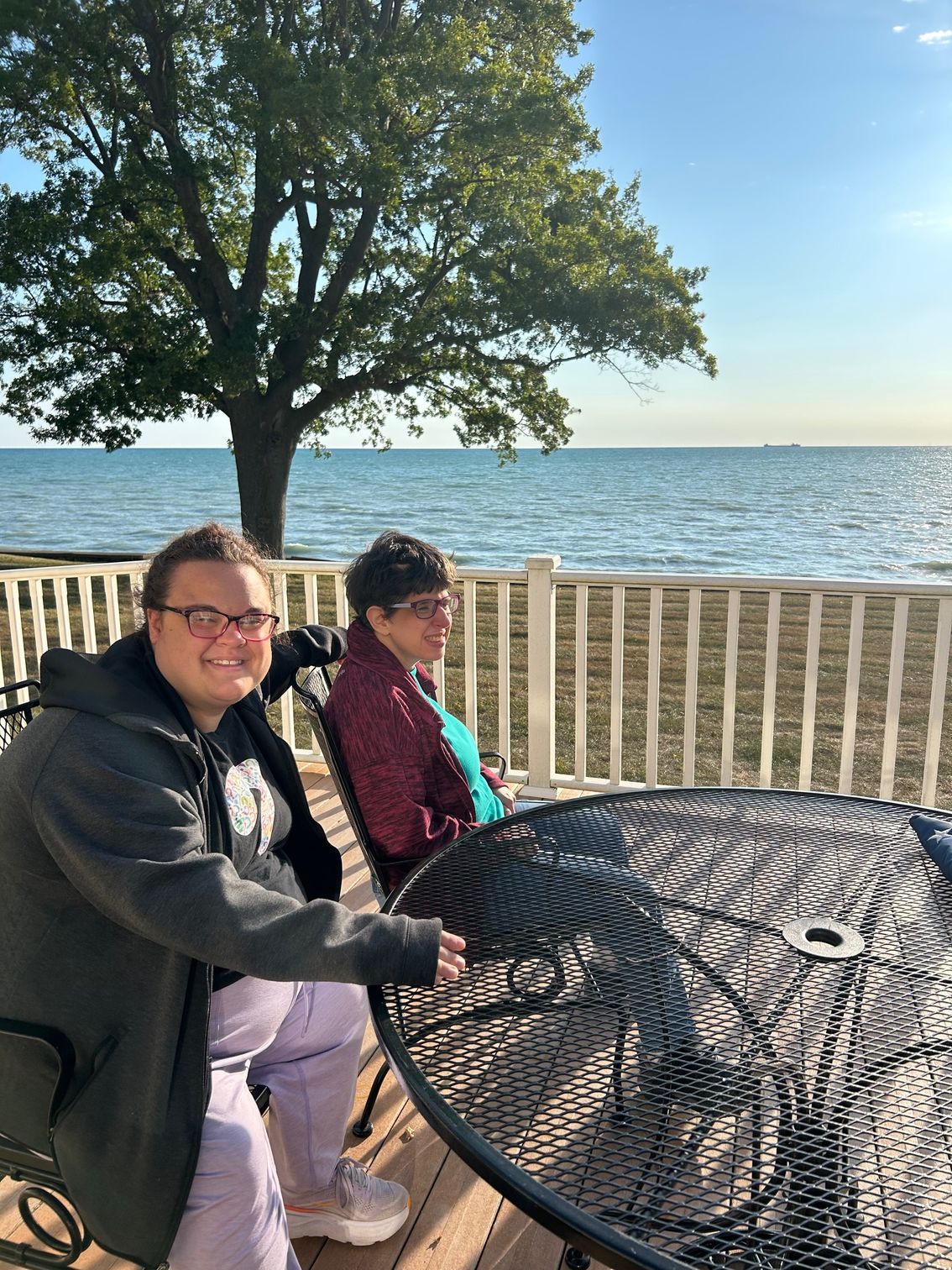 Two people sit at a round table, facing the camera near a body of water under a tree.