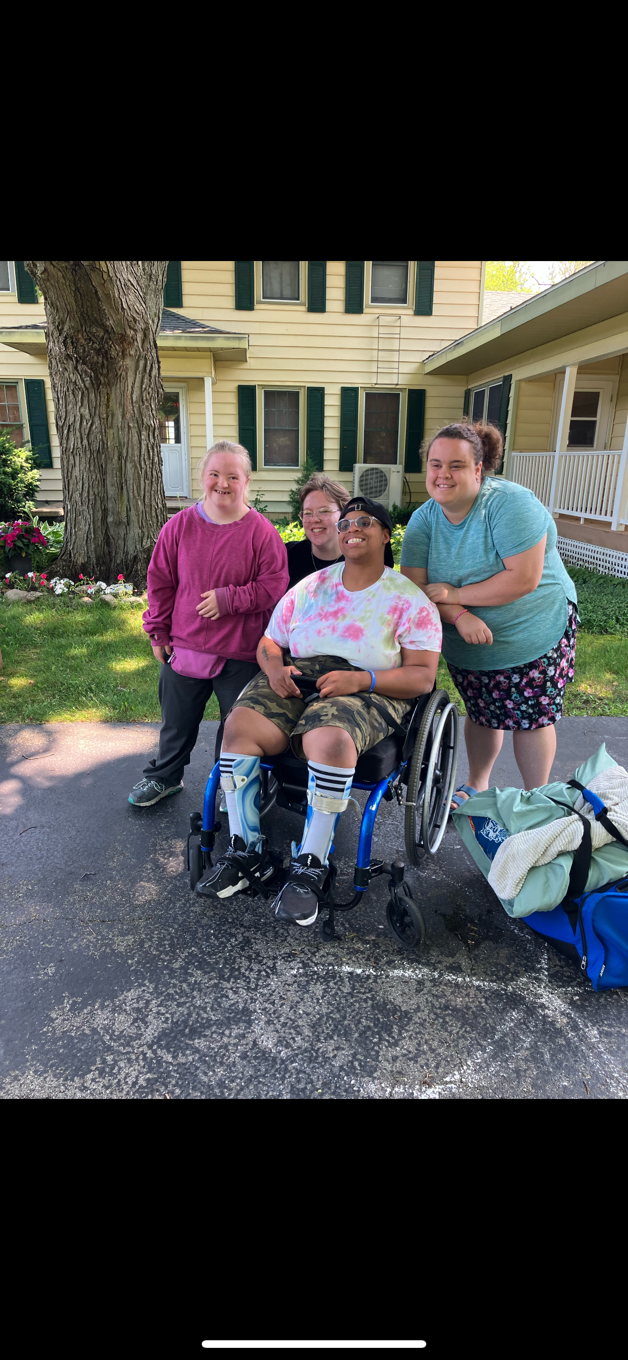 Four people posing outdoors, one in a wheelchair. A house with shutters is behind them.