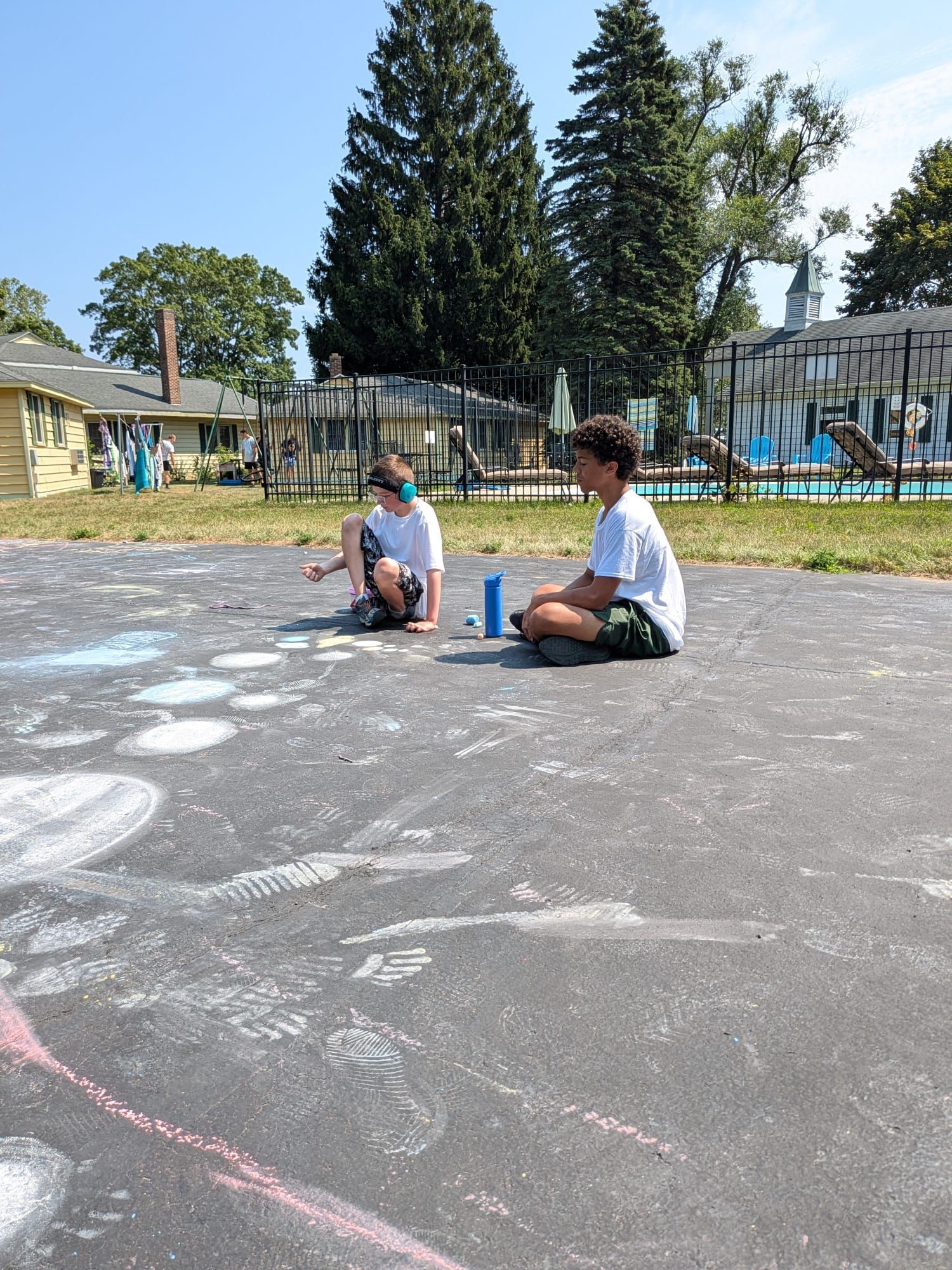 Two people seated on asphalt, near chalk drawings. Blue container sits between them; trees in the background.