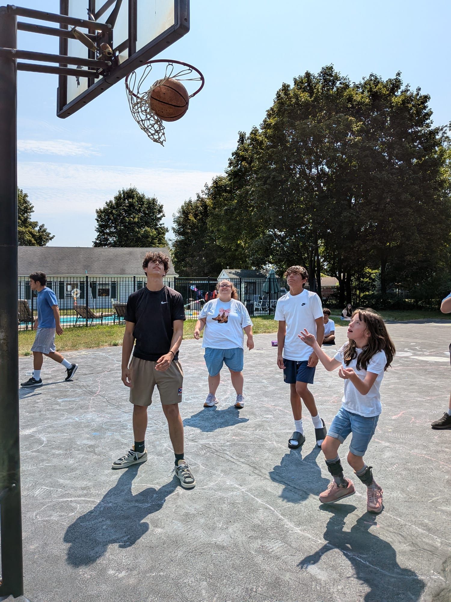 People playing basketball outdoors, with one person under the hoop and others watching nearby.