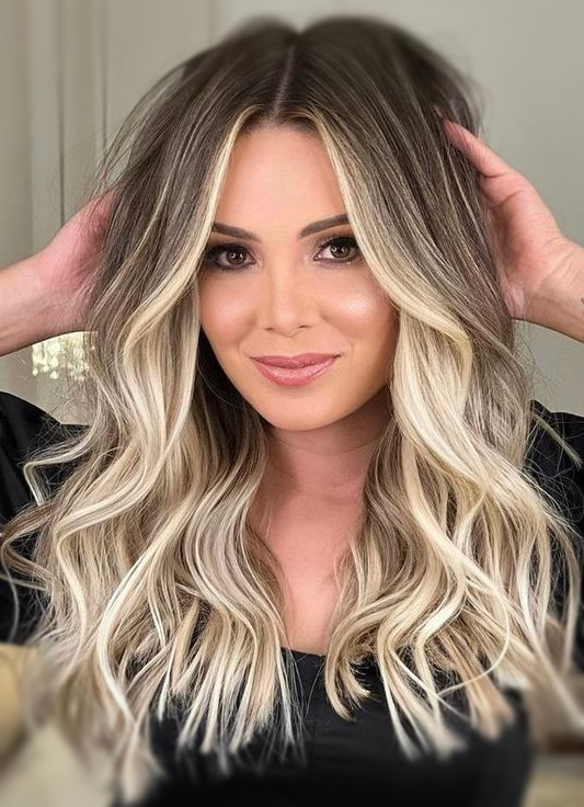 Donna con capelli biondi e castani ondulati, mani vicino alla testa, sorridente.
