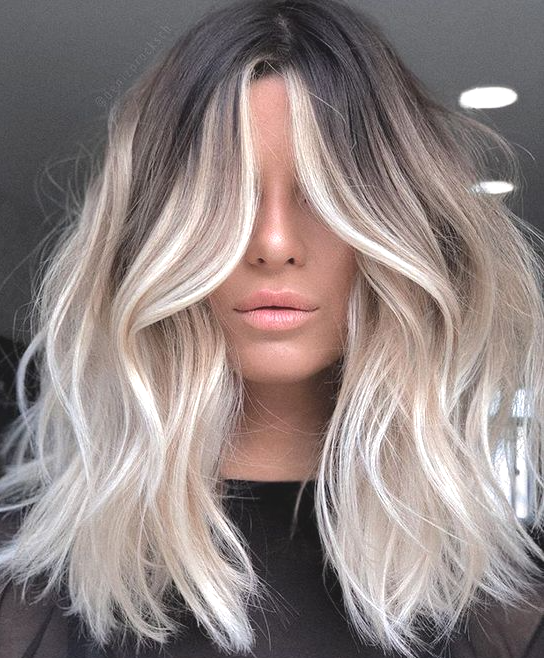 Donna con radici scure e mèches bionde che incorniciano il viso, capelli ondulati lunghi fino alle spalle.