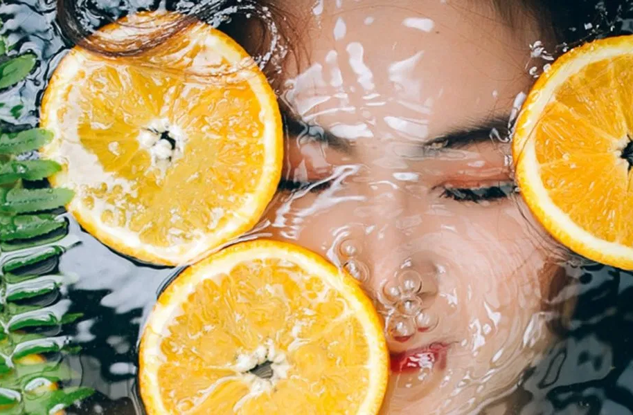Volto di donna immerso nell'acqua, circondato da fette d'arancia e fogliame verde. Gocce d'acqua visibili.