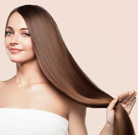 Donna con lunghi capelli castani lisci, sorridente, che tiene i capelli con la mano destra. Sfondo beige.