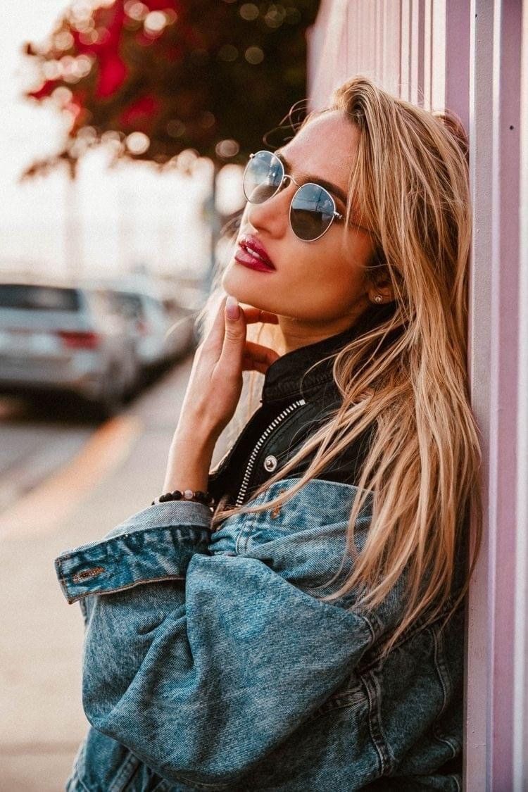 Una donna con gli occhiali da sole è appoggiata a un muro rosa, indossa una giacca di jeans e si tocca il viso.