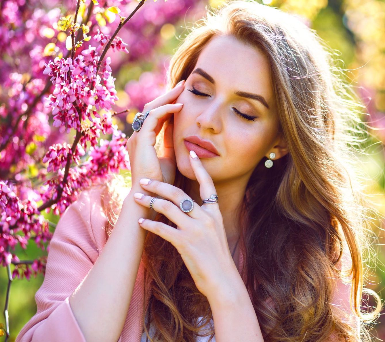Donna con gli occhi chiusi vicino a fiori rosa, che si tocca il viso. Capelli castano chiaro, gioielli, indossa una giacca rosa.