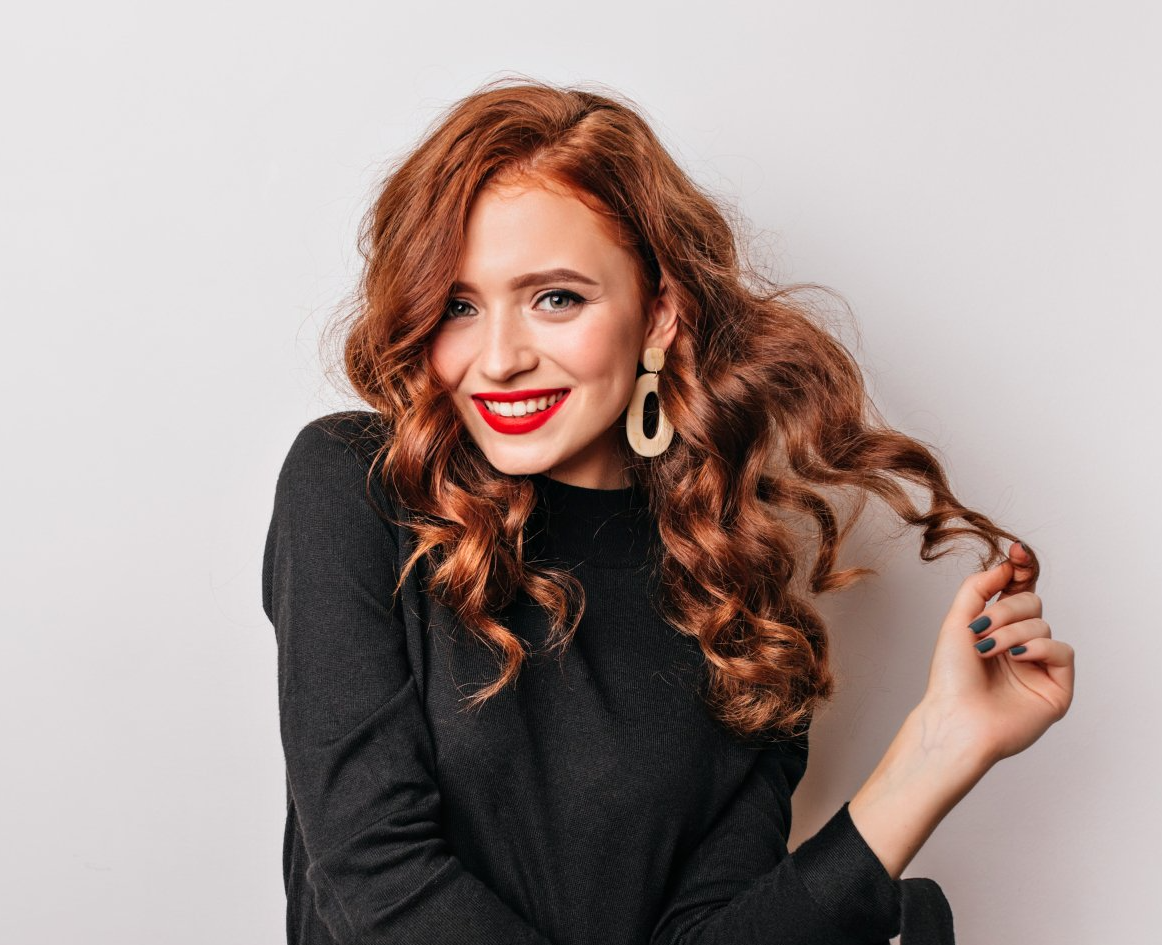 Una donna con i capelli rossi e ricci sorride e si tocca i capelli. Indossa un top nero, rossetto rosso e orecchini su uno sfondo bianco.