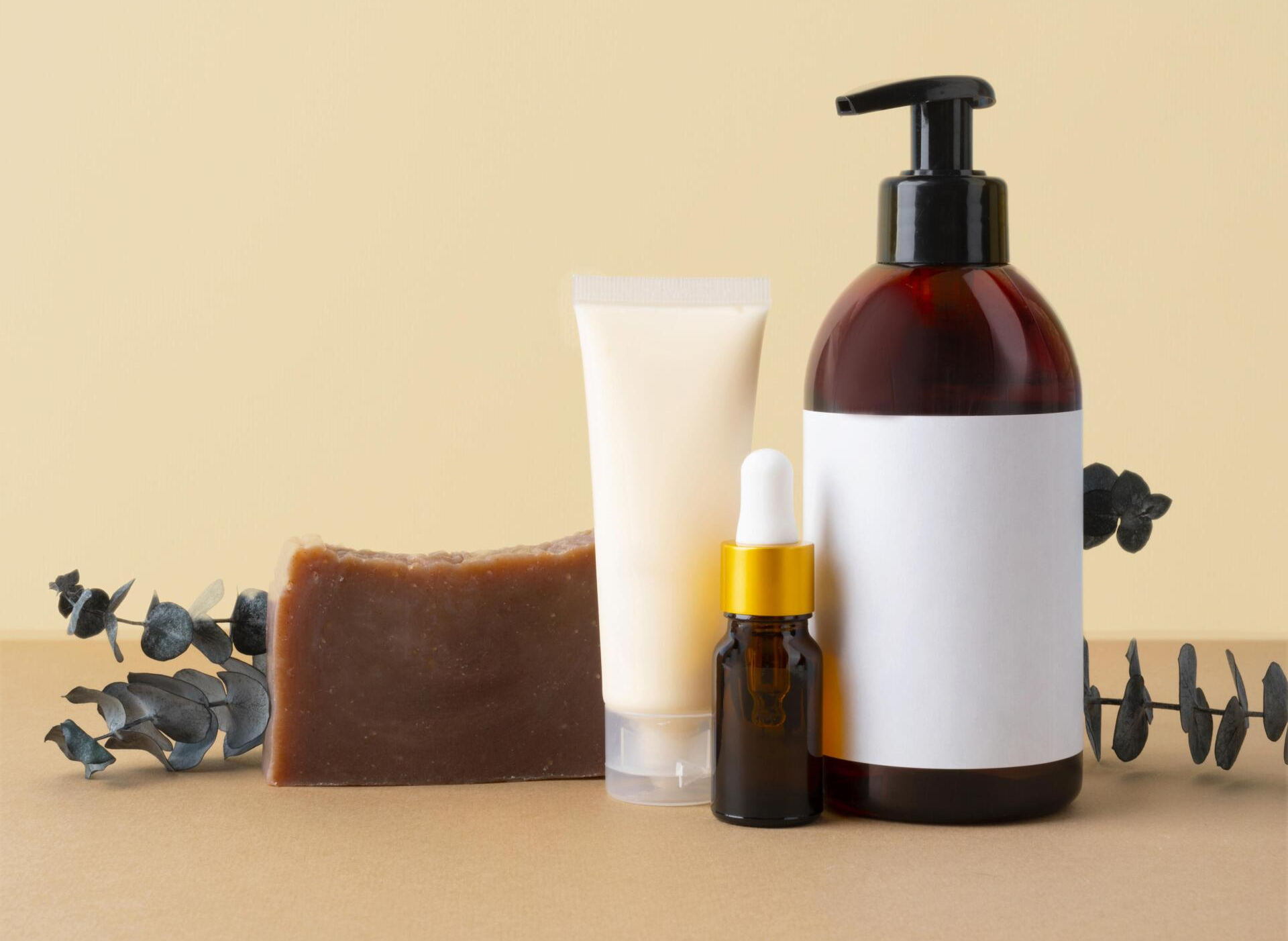 Prodotti di bellezza disposti su una superficie beige: sapone, tubetto di crema, olio essenziale e flacone con pompa e etichetta vuota.