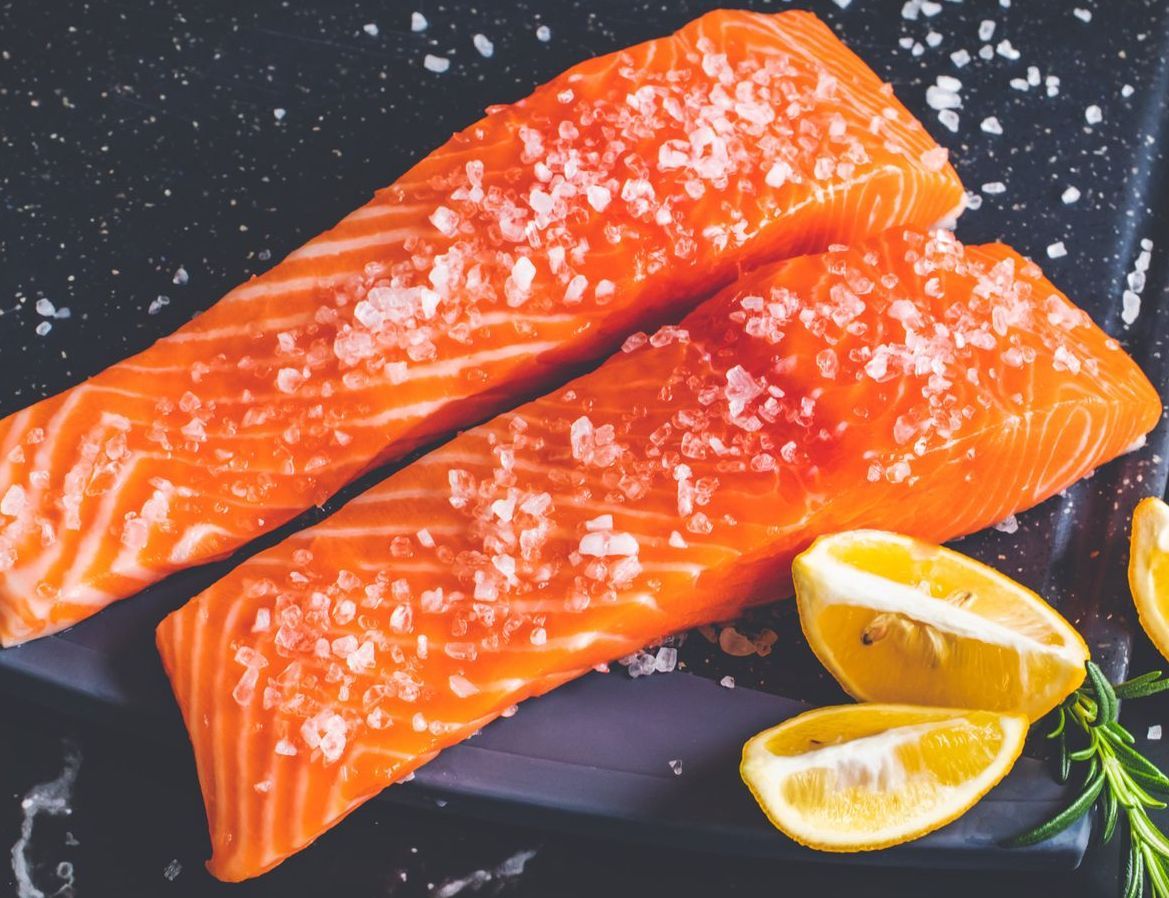 Due filetti di salmone crudo cosparsi di sale, spicchi di limone e rosmarino su una superficie scura.