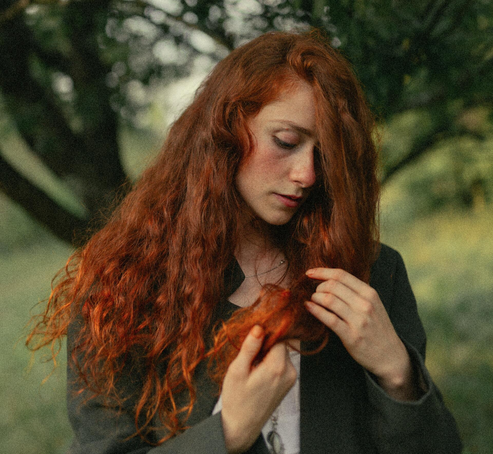 Donna con lunghi capelli rossi e ondulati in un ambiente esterno sfocato, che si tocca una ciocca di capelli.