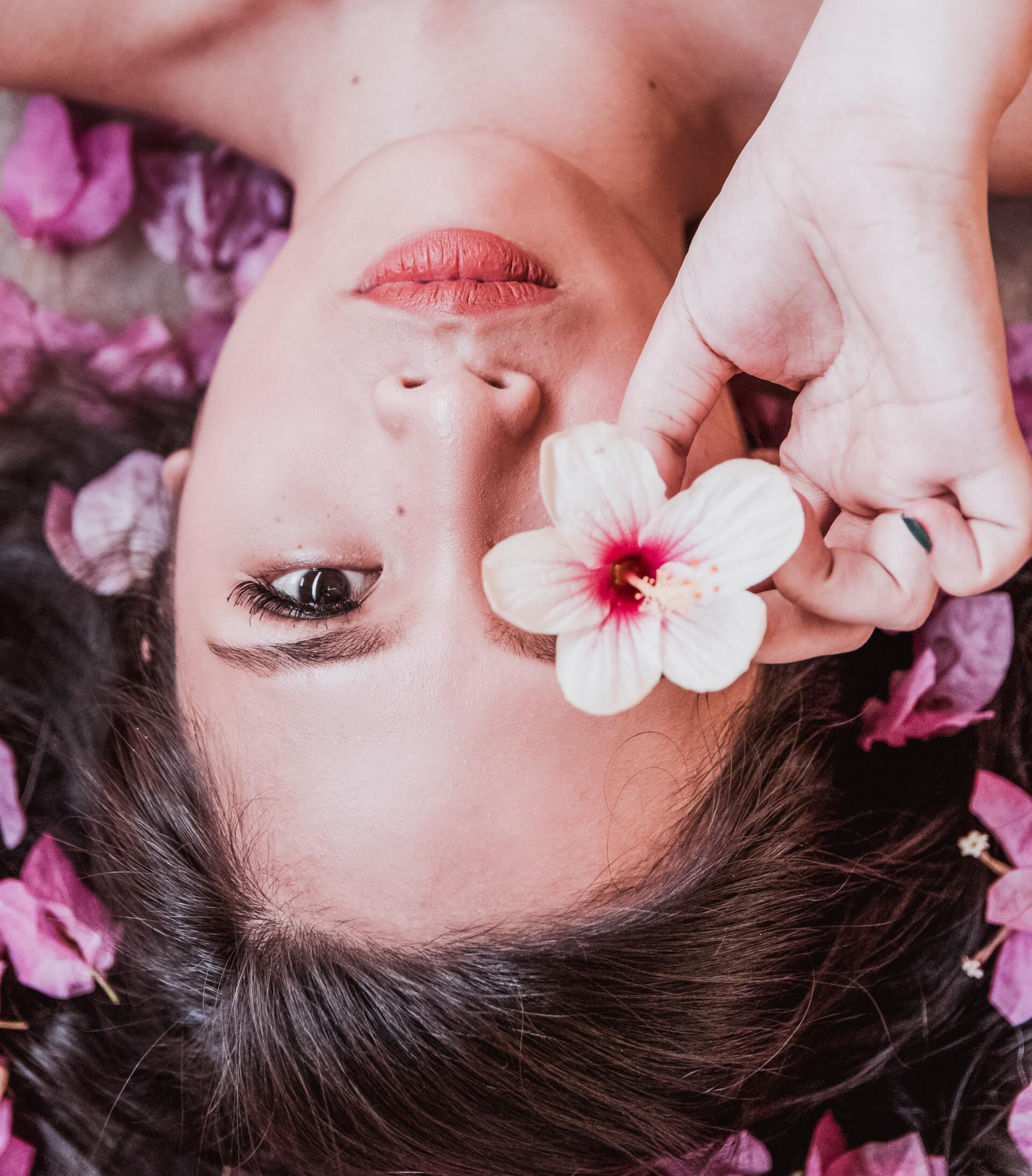 Donna con i capelli scuri che guarda la telecamera mentre tiene un fiore bianco sopra l'occhio, circondata da fiori rosa.